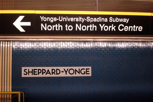 Sign on blue tile wall reads SHEPPARD-YONGE on a metal plate. Overhead sign in ‘Helvetica’ reads &larr; Yonge-University-Spadina Subway North to North York Centre